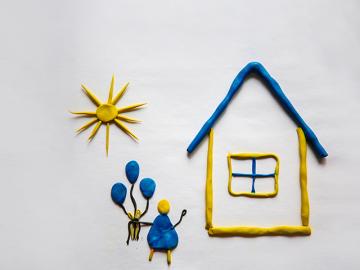 Yellow house with a blue roof under a yellow sun. a child in blue clothes stands nearby and holds a bunch of blue balloons