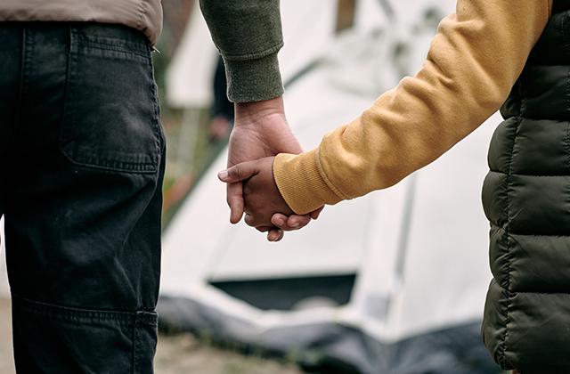 Parent of elder brother holding hand of child against tent in migrant camp